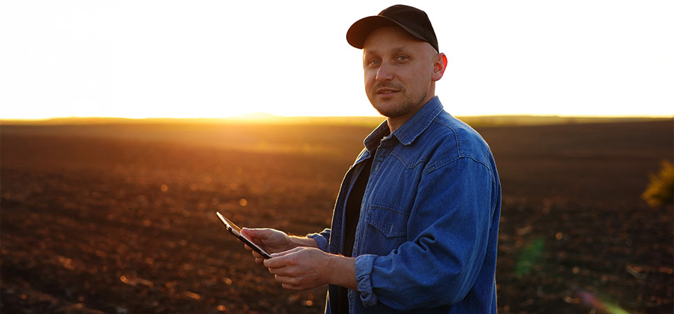 Agricultor segurando um tablet no campo ao pôr do sol, representando o uso de tecnologia como drones e satélites para monitoramento agrícola